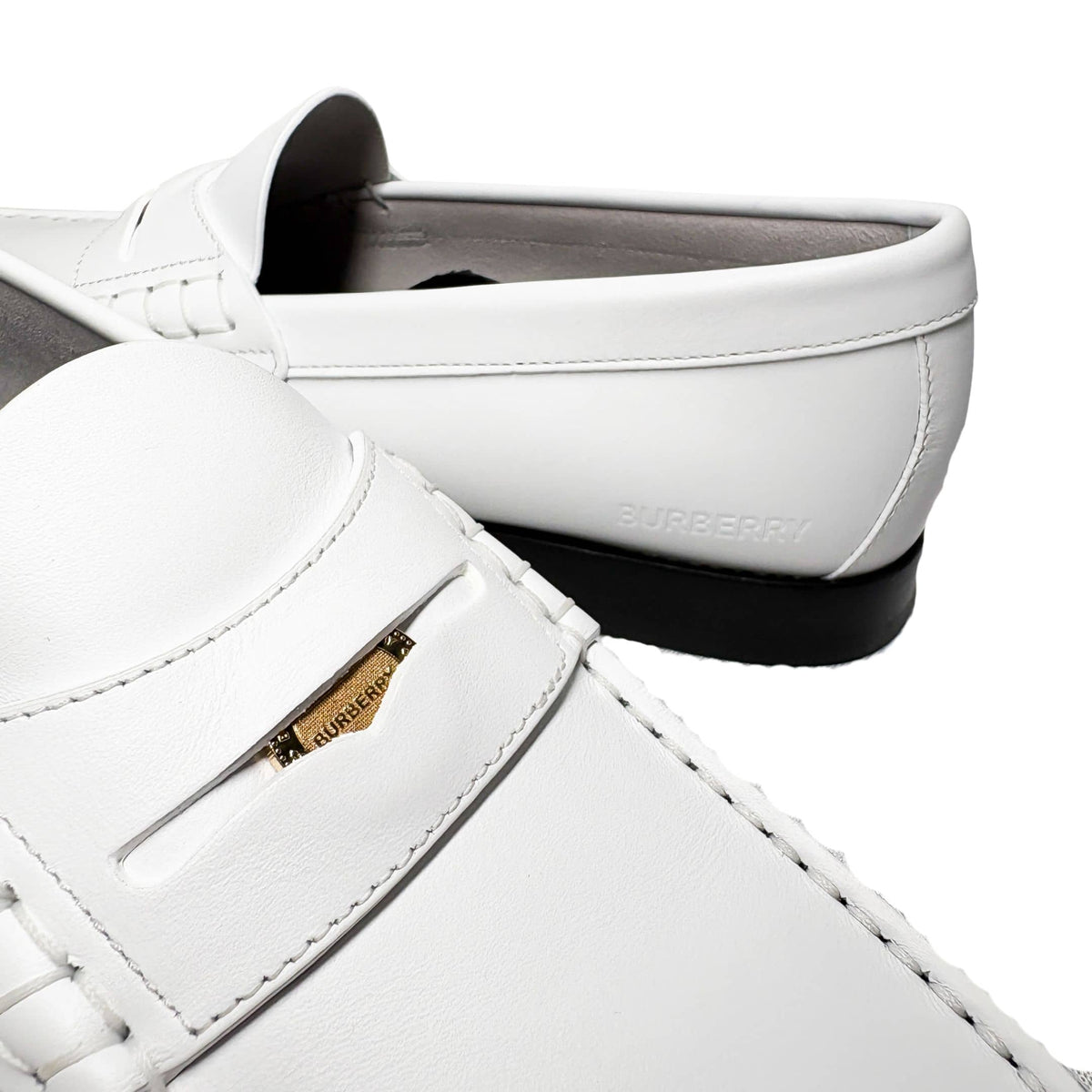Asymmetrical studio layout of the white leather Burberry Rupert Coin Loafers, with one shoe angled forward to display the side profile.