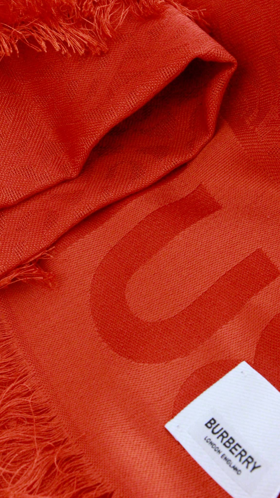 Close-up view of the vermillion red Burberry TB monogram jacquard scarf showing fabric weave and white logo care tag.