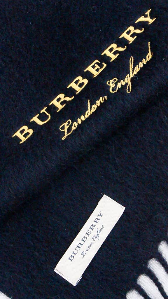 Angled close-up of black Burberry cashmere scarf showing yellow gold embroidered logo and white designer tag.