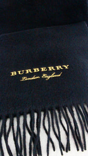 Frontal close-up view of the black Burberry cashmere scarf highlighting the metallic gold logo embroidery and fringed hem.