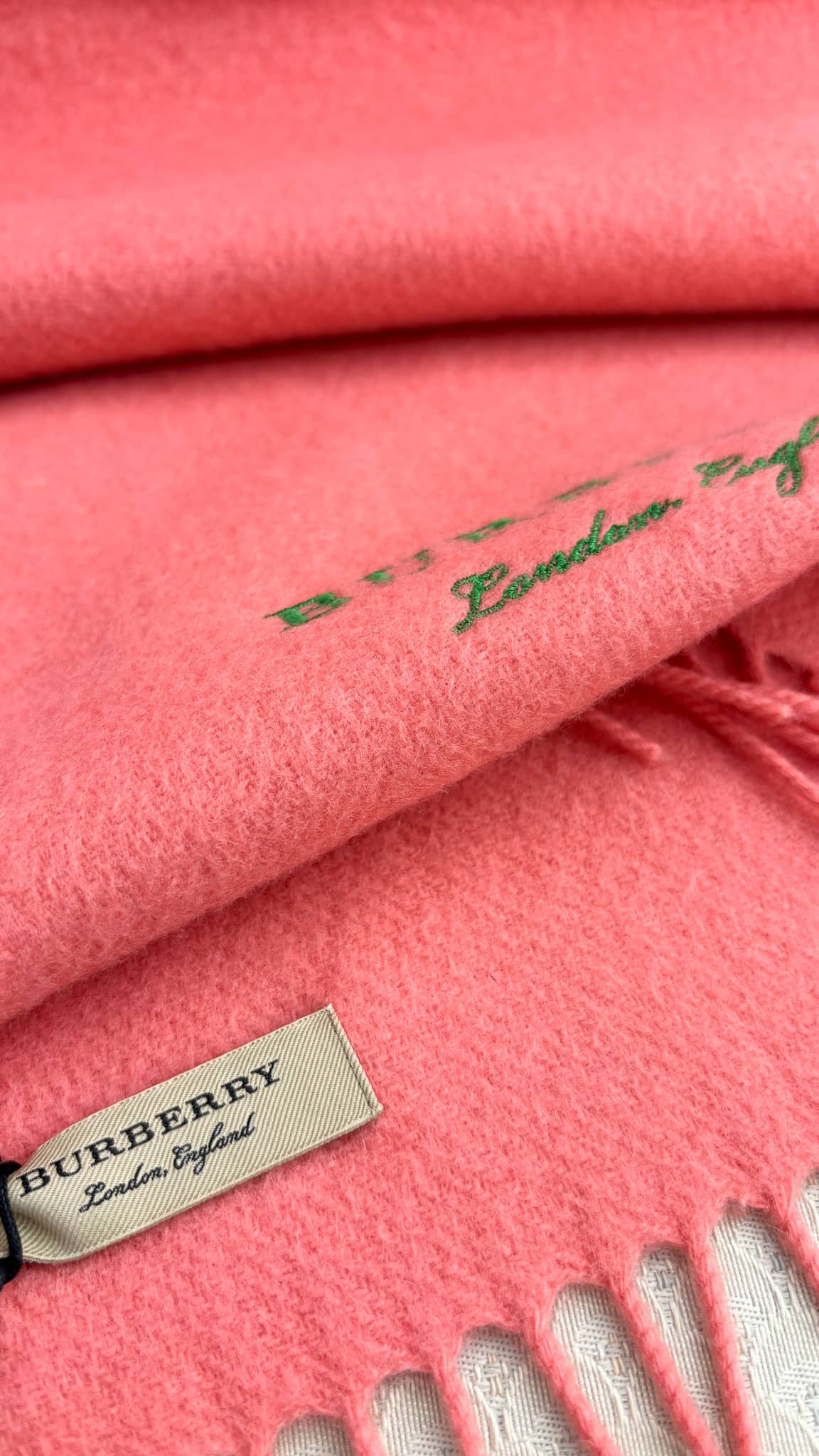 Close-up view of the coral Burberry cashmere scarf displaying the official brand woven label and fringed edge.