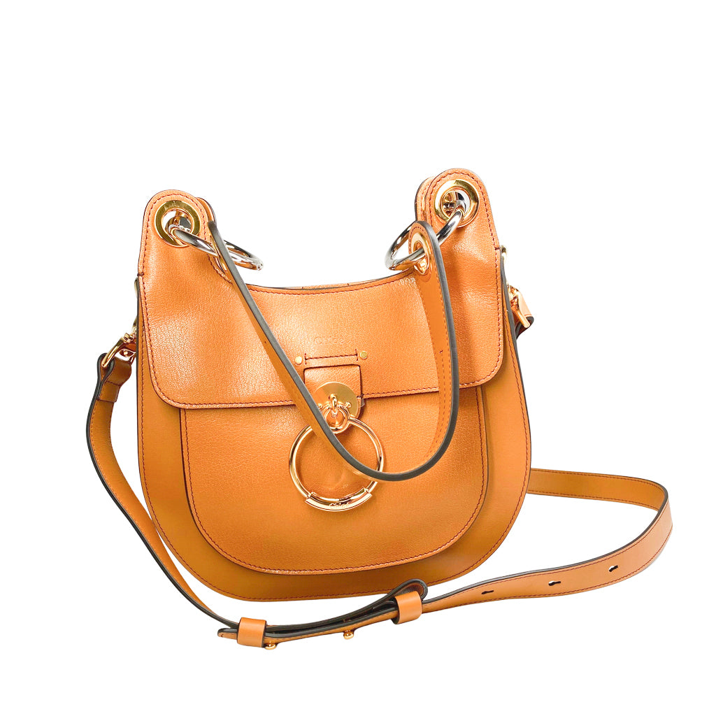 Front studio view of the Chloé Tess shoulder bag in tan leather against a white background, featuring its curved saddle shape and long crossbody strap.