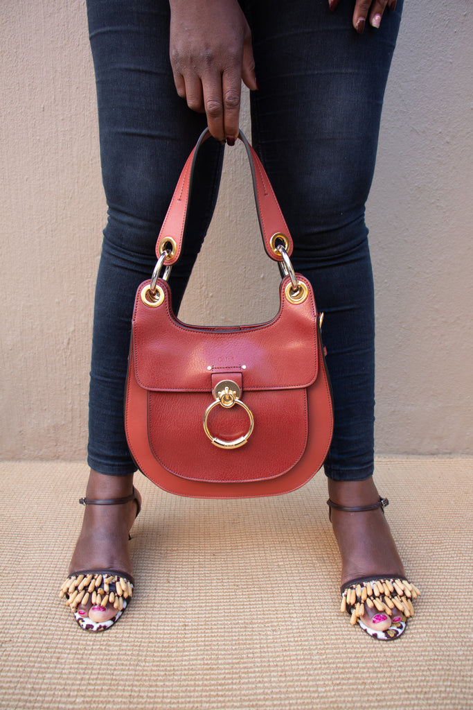 On-foot wearing view of a model carrying the red leather Chloé Tess bag by its short top handle strap, styled with dark denim jeans.