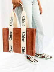 Side profile view of a model carrying the brown suede Chloé Woody tote bag over the shoulder, paired with patterned loafers.