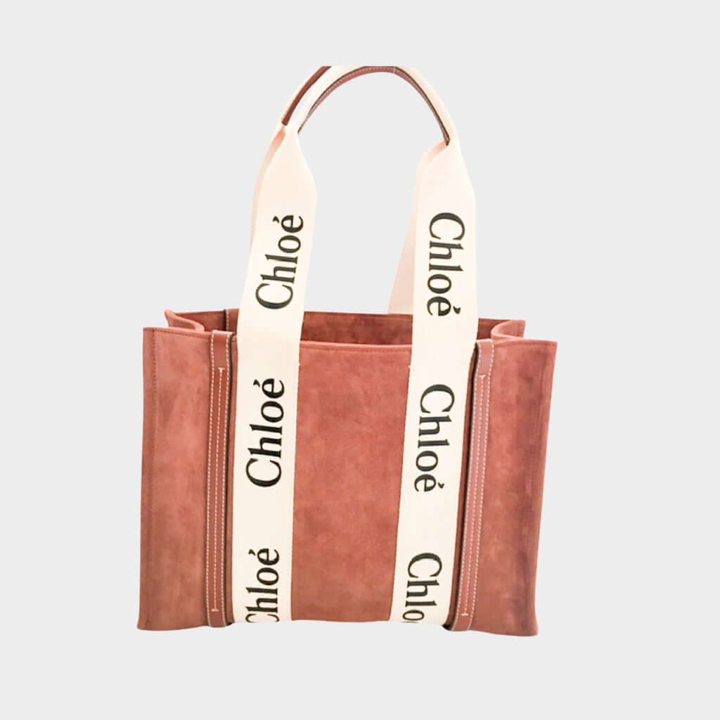 Front view of the Chloé Woody Medium Tote Bag in brown suede, highlighting the cream canvas logo-printed shoulder straps and smooth brown leather trim.