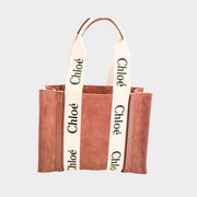 Front view of the Chloé Woody Medium Tote Bag in brown suede, highlighting the cream canvas logo-printed shoulder straps and smooth brown leather trim.
