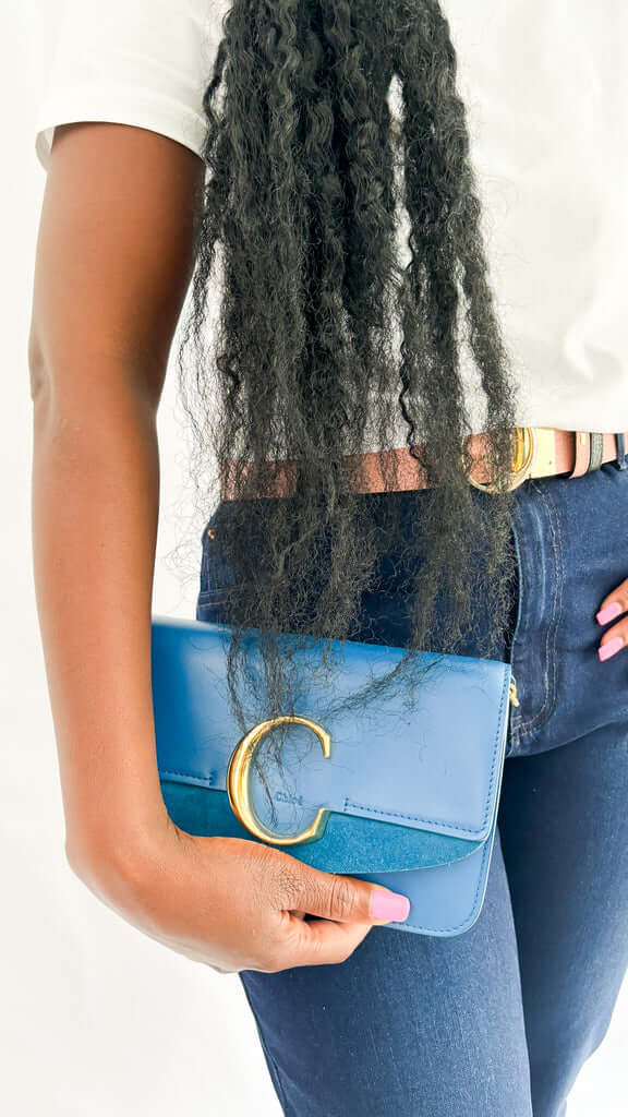 On-foot wearing view of a model carrying the blue leather Chloé C handbag as a clutch, styled with dark denim jeans and a leather Gucci belt.
