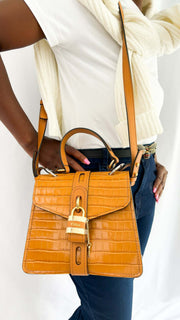 On-foot wearing view of a model carrying the Chloé Aby Day brown embossed leather handbag by its top handle, paired with a classic Gucci belt.