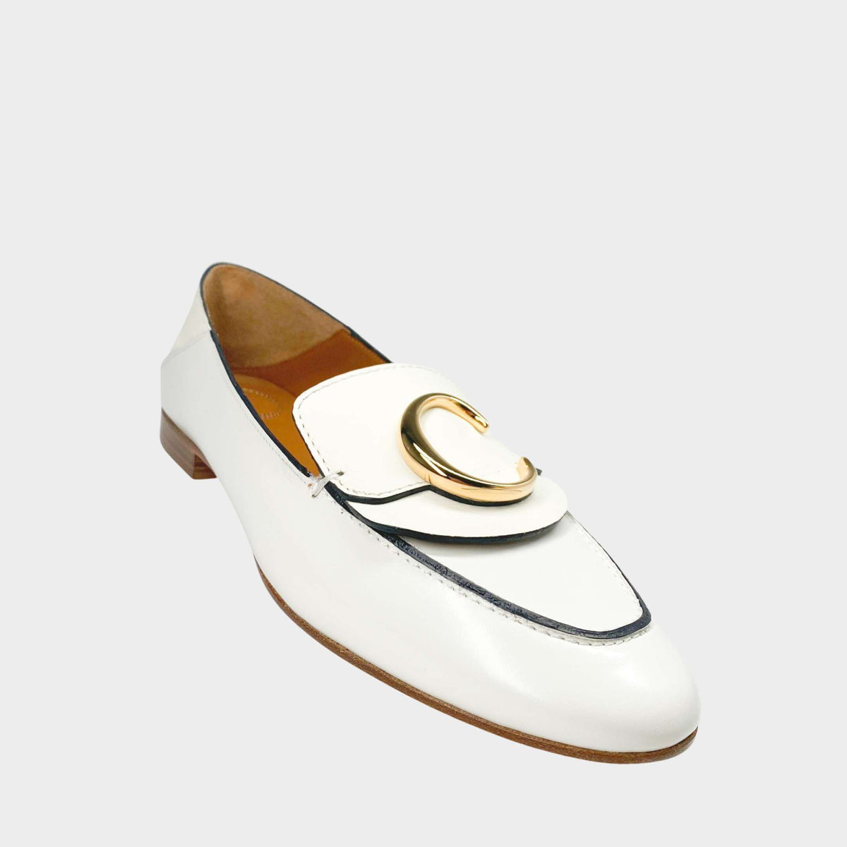 Side profile close-up of a Chloé C loafer in white patent leather, showcasing the prominent gold-toned C initial logo on the strap and the stacked flat heel.