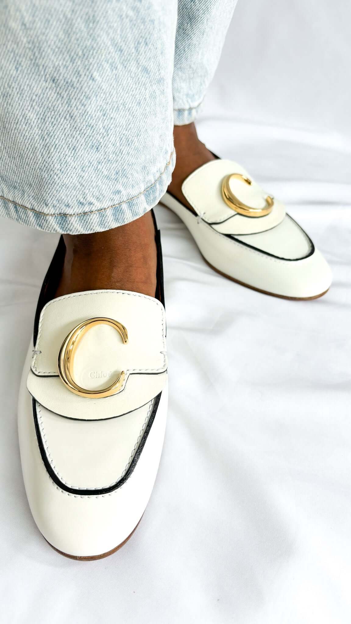 On-foot wearing view of a pair of white patent leather Chloé C loafers styled with light wash denim jeans, highlighting the round toe design.