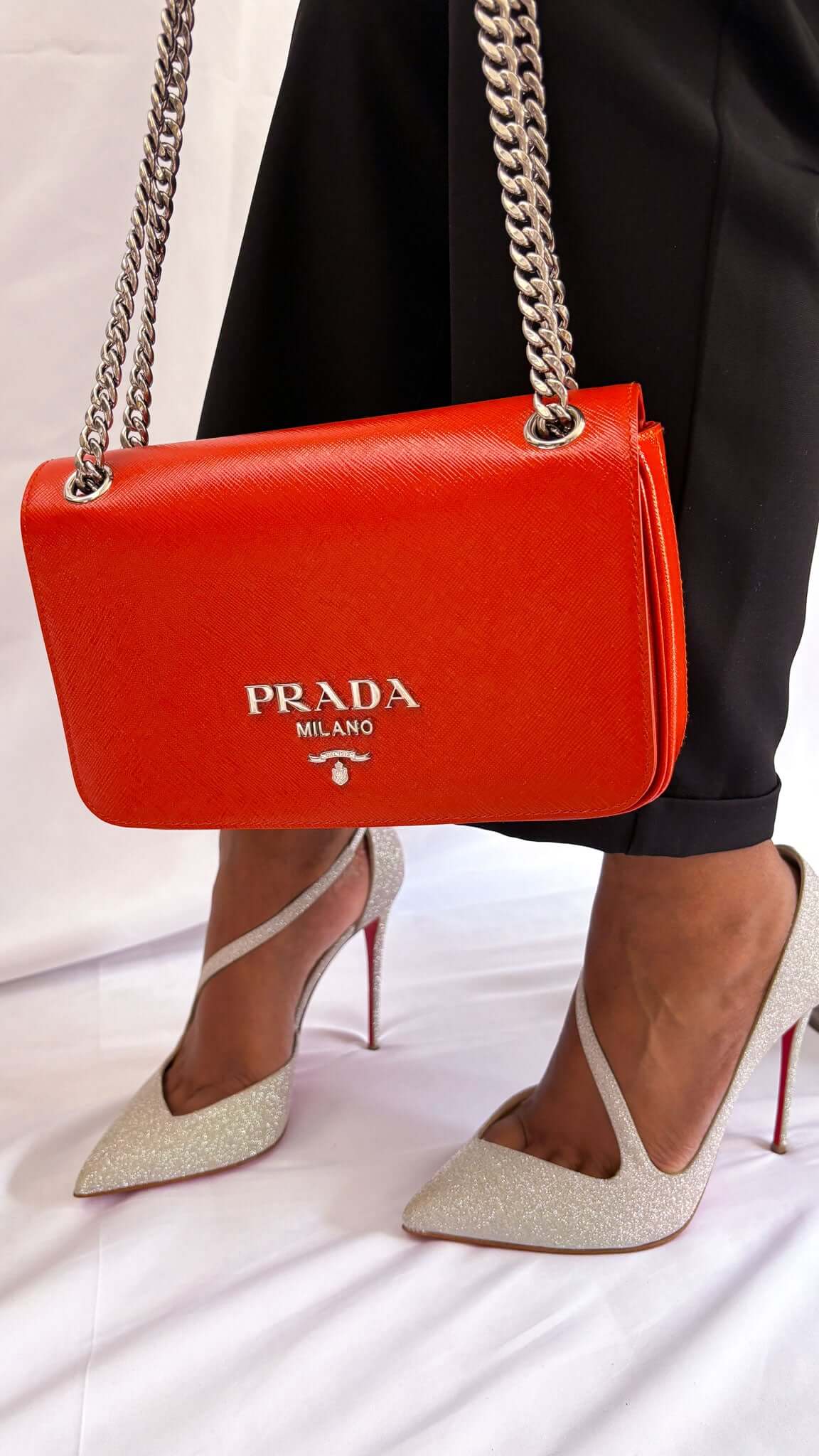 On-foot wearing view of a model in Christian Louboutin silver glitter pumps, carrying a vibrant red Prada shoulder bag with a silver chain strap.