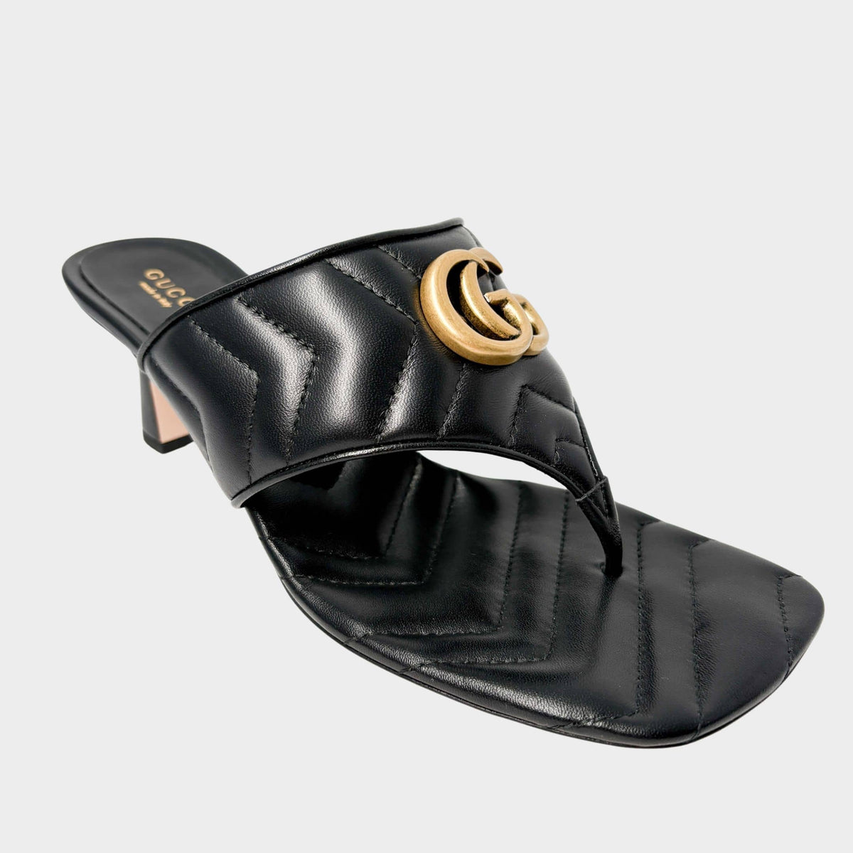 Side profile view of a Gucci Charlotte thong sandal in premium black quilted leather, featuring a prominent gold-toned GG Marmont logo and a comfortable low kitten heel.
