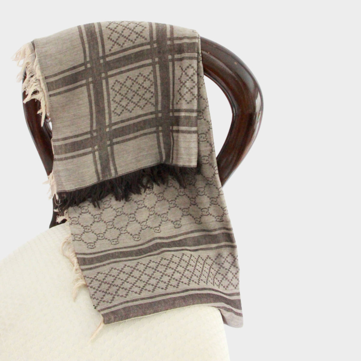 Gucci GG monogram scarf in brown wool draped elegantly over a chair back showing plaid pattern and fringe trim.