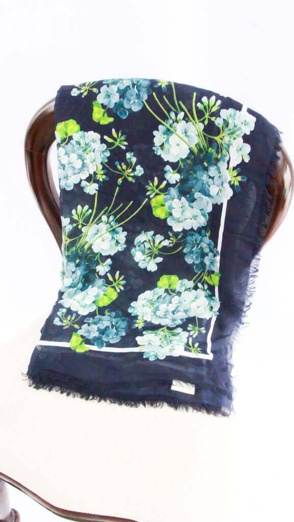 Front view of the Gucci Bloom silk scarf in navy blue, featuring vibrant blue and white floral graphics with green foliage draped on a display mannequin.