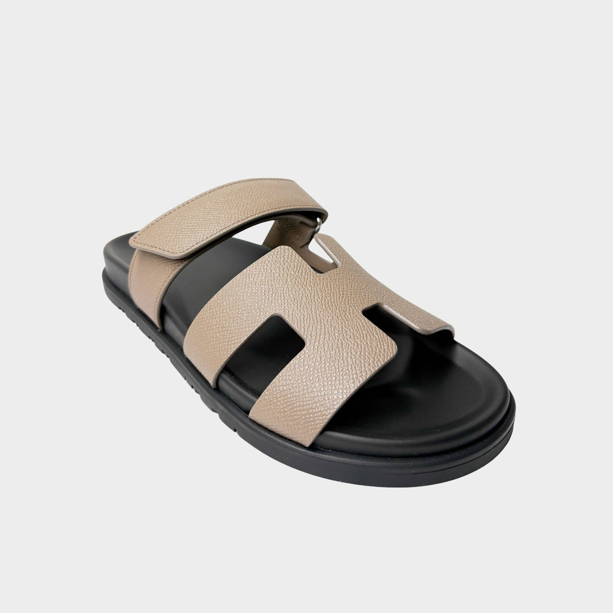Side profile view of an Hermès Chypre sandal in Etoupe taupe calfskin leather, showcasing the iconic H-shaped upper strap and adjustable velcro top strap.
