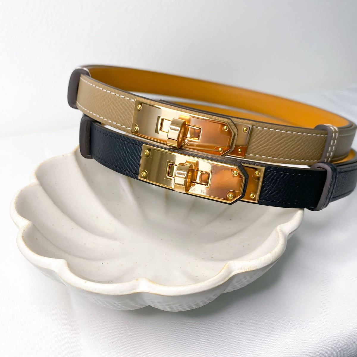Hermès Kelly belts in Étoupe, navy blue, and gold leather with rose gold buckles displayed on decorative dish