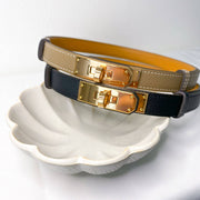 Hermès Kelly belts in Étoupe, navy blue, and gold leather with rose gold buckles displayed on decorative dish