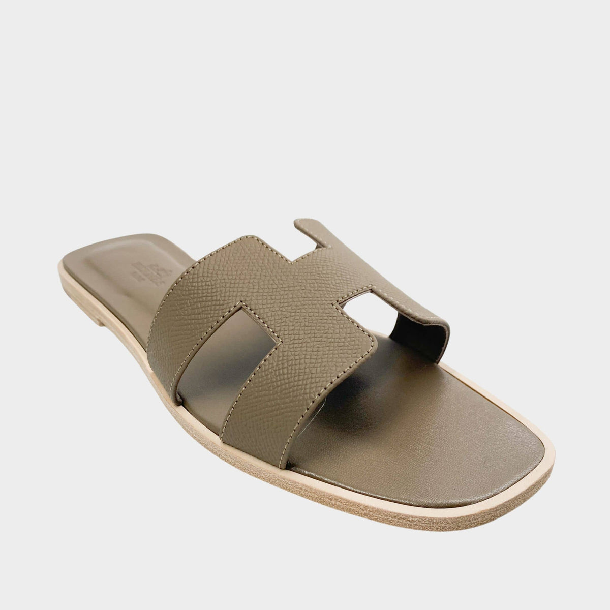 A crisp product shot capturing a single classic Hermès Oran slide tilted slightly to reveal the side edge paint and the seamless connection of the iconic H leather flap to the slim sole.
