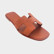 A striking side profile shot of the Hermès Oran slide, showcasing the vibrant Orange Canyon leather upper and the seamless, low-profile sole construction.