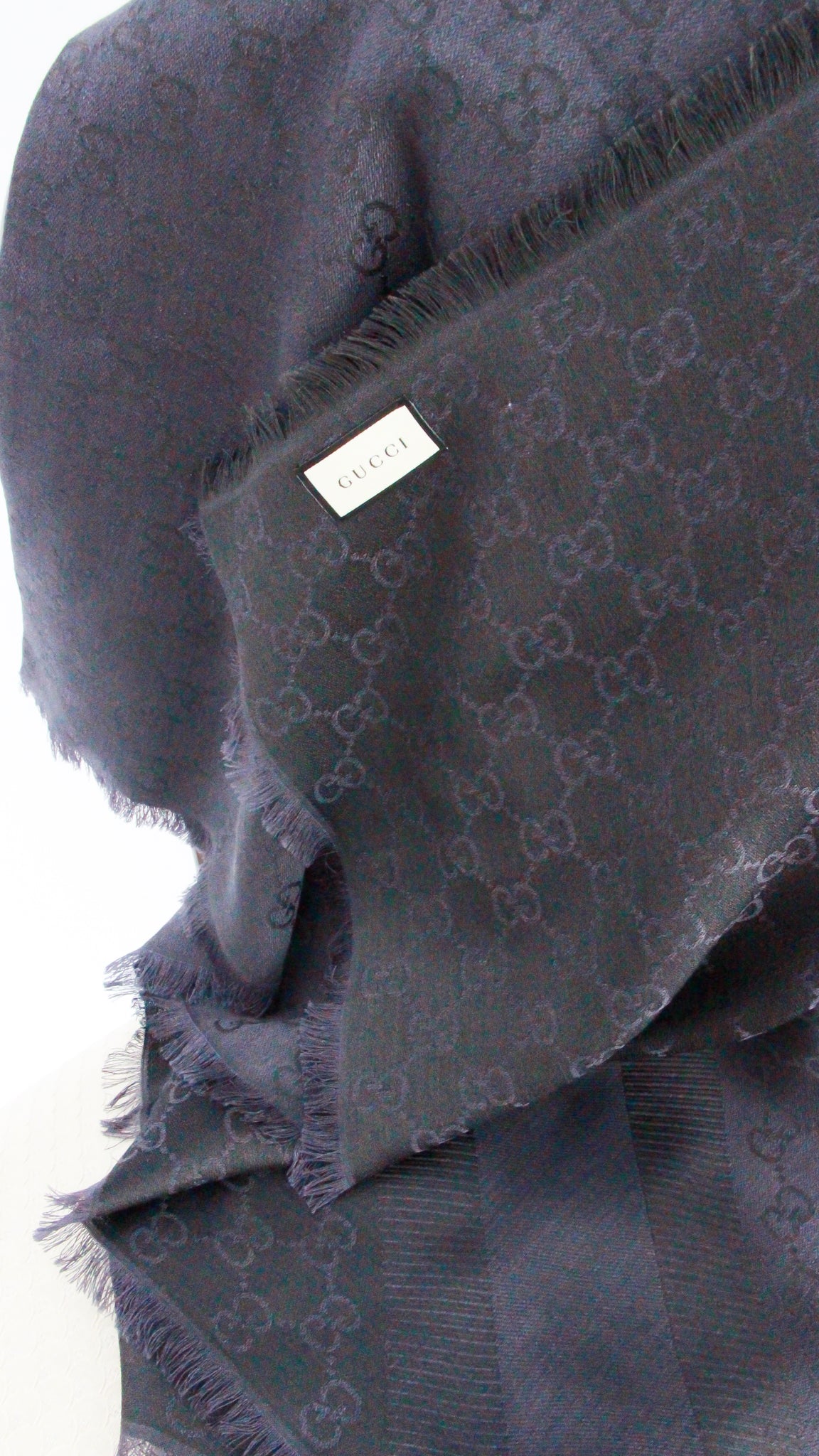 Close-up view of woven GG pattern, fabric care label, and frayed fringe border on navy Gucci scarf.