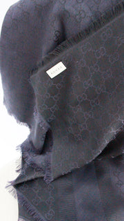 Close-up view of woven GG pattern, fabric care label, and frayed fringe border on navy Gucci scarf.