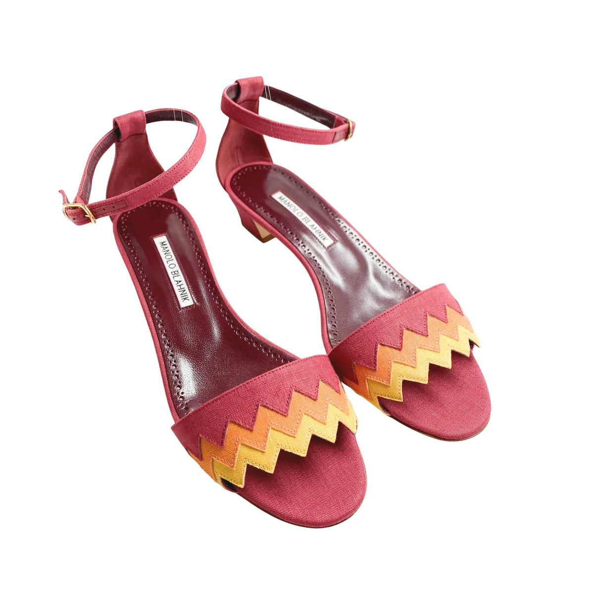 Three-quarter studio view of a pair of Manolo Blahnik Lochpri canvas ankle-strap sandals displaying yellow and orange zig-zag vamp details.