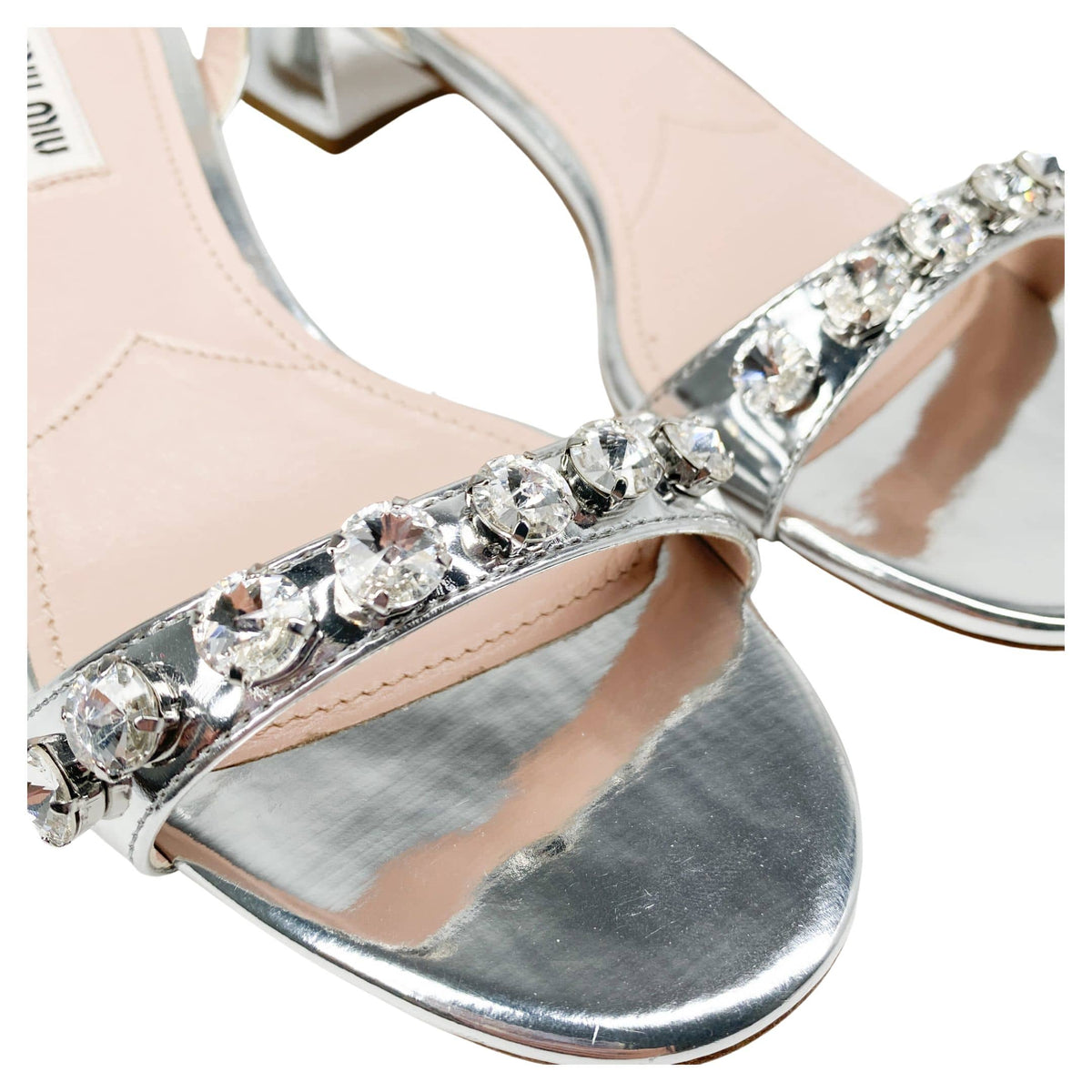 Close-up macro view of shimmering crystal embellishments across the front strap of Miu Miu silver leather sandals.