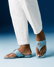 On-foot wearing view of the Gucci Marmont Charlotte sandals in Foggy Sky, styled with white linen trousers in a photo studio setup.