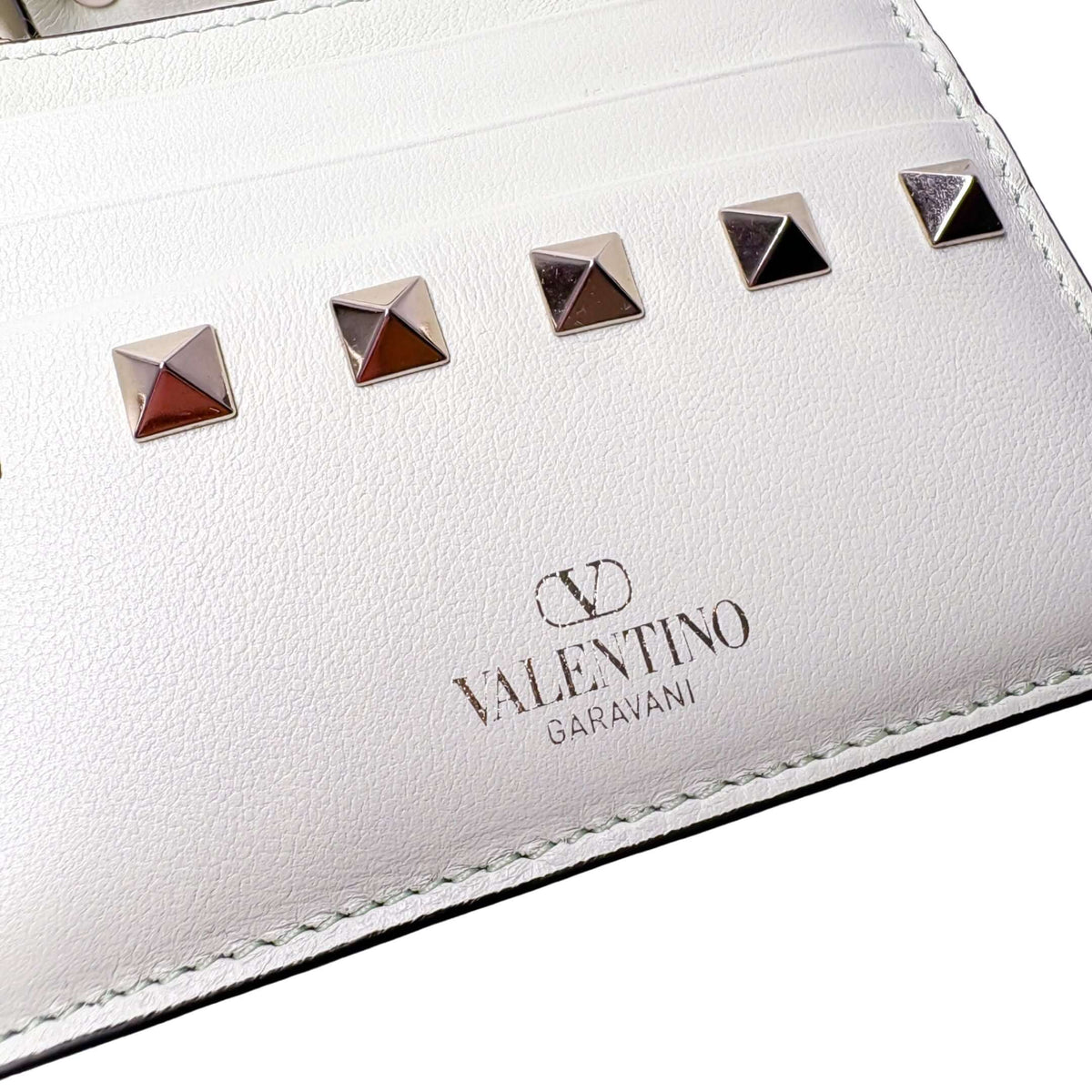 Close-up of the white Valentino card holder, showcasing the gold embossed logo and polished rockstud embellishments.