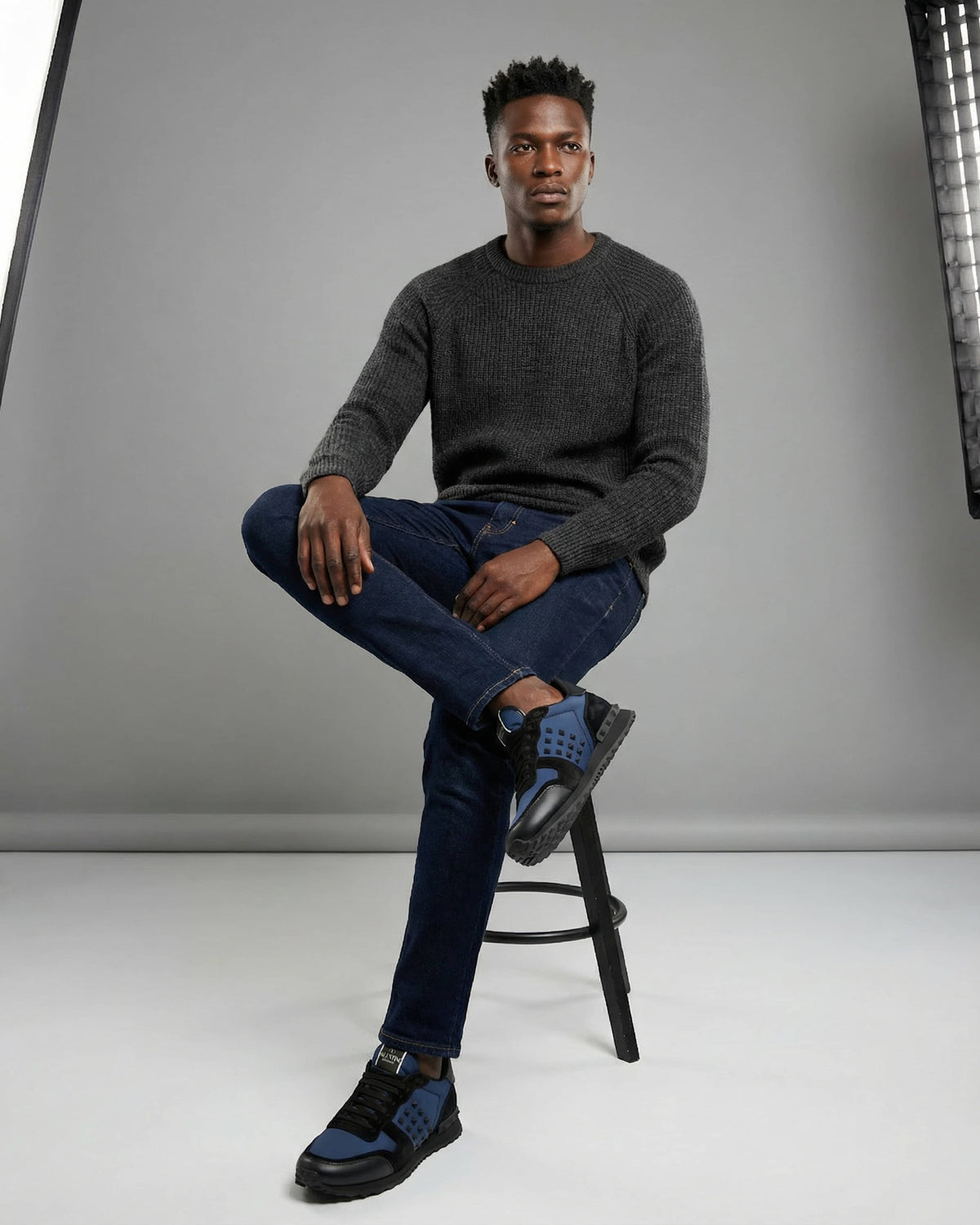 Full-length model view wearing navy Valentino Rockrunner sneakers seated in a grey studio setting.